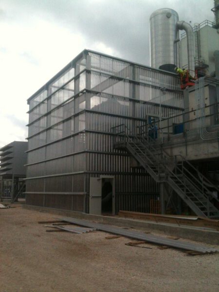 A large industrial structure with metal exterior, stairs on the side, and a cylindrical tower on top, under a cloudy sky.