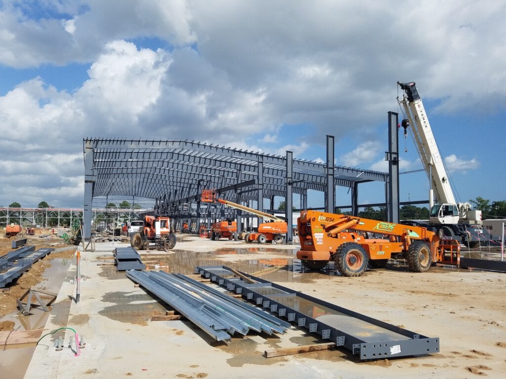 Construction site with steel frames being erected. Multiple orange construction vehicles and cranes are visible under a partly cloudy sky.