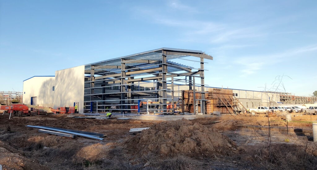 Construction site with a large steel frame building in progress, surrounded by dirt and construction materials under a clear sky.