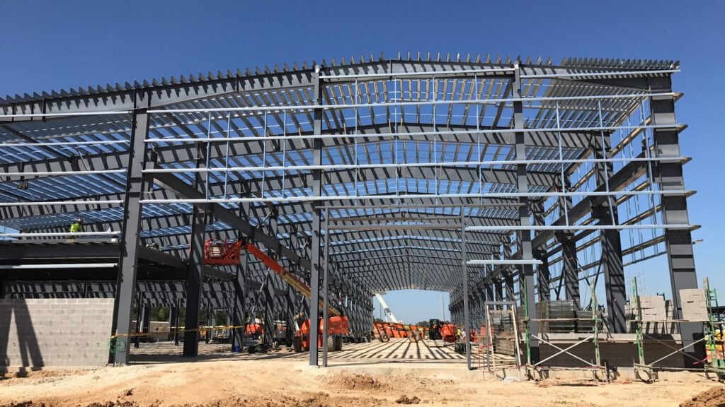 Large steel-frame structure under construction, with cranes and construction materials on site, against a clear blue sky.