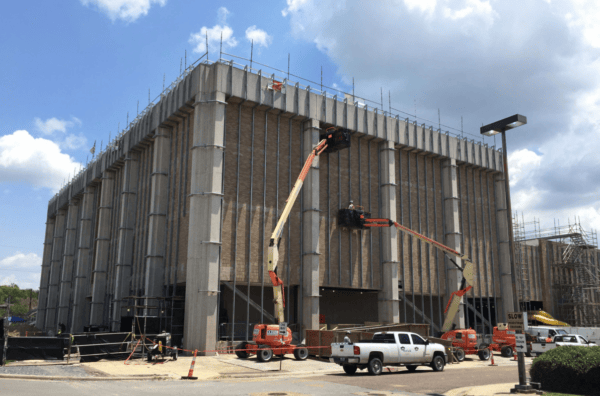 A large building under construction with cranes, scaffolding, and workers. Several vehicles are parked nearby. The sky is partly cloudy.