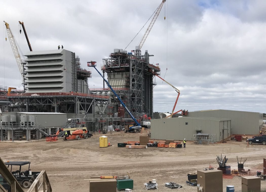 Construction site with cranes, unfinished steel structures, and machinery. Workers in hard hats are visible among equipment and stacked materials.