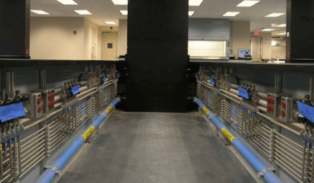 A hallway with metallic pipes and blue tubes along the sides, leading to a black structure in an industrial construction room with ceiling lights.