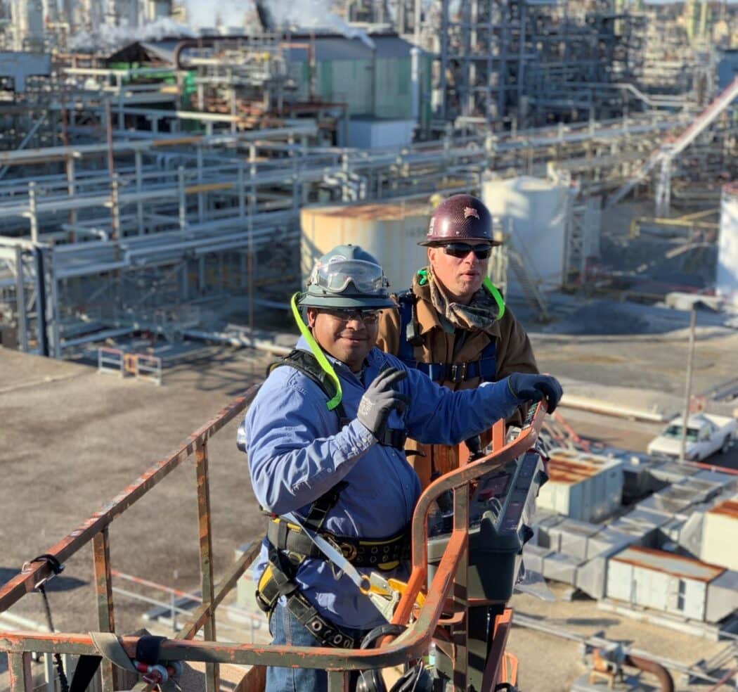 A construction worker in safety gear, including a helmet and harness, stands on scaffolding next to a building with large windows.