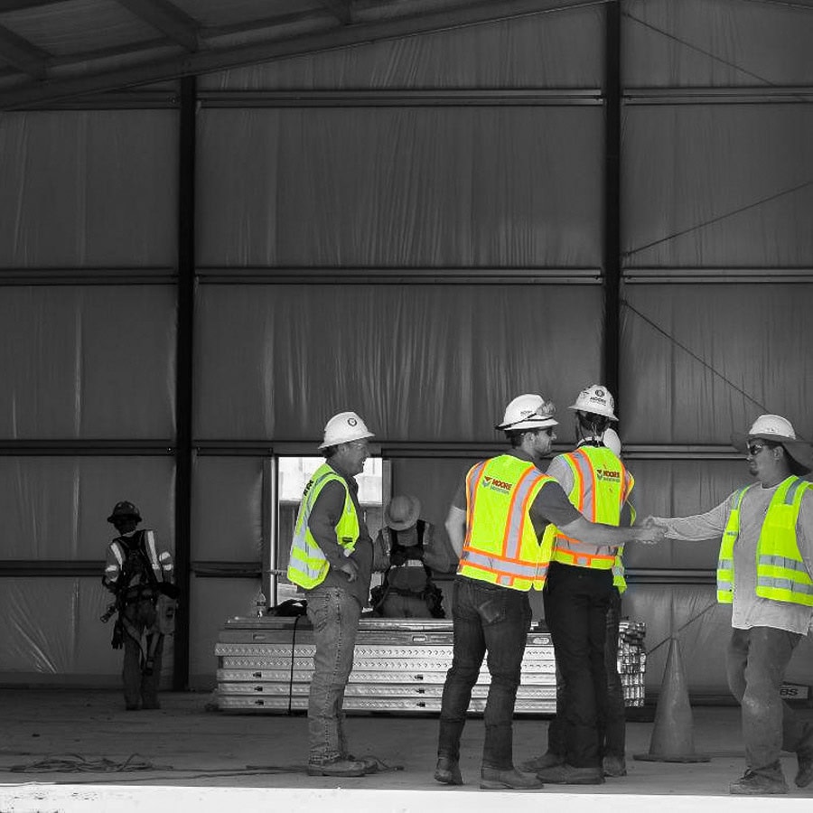 Workers in safety vests and hard hats stand inside a building under construction, conversing. Metal beams and construction equipment are visible.