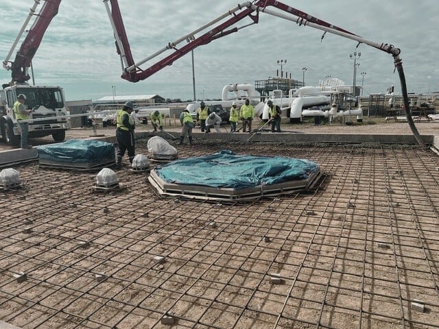 Construction site poised for a concrete pour; workers prep over a steel rebar grid while machinery awaits under a blue tarp for system replacement.