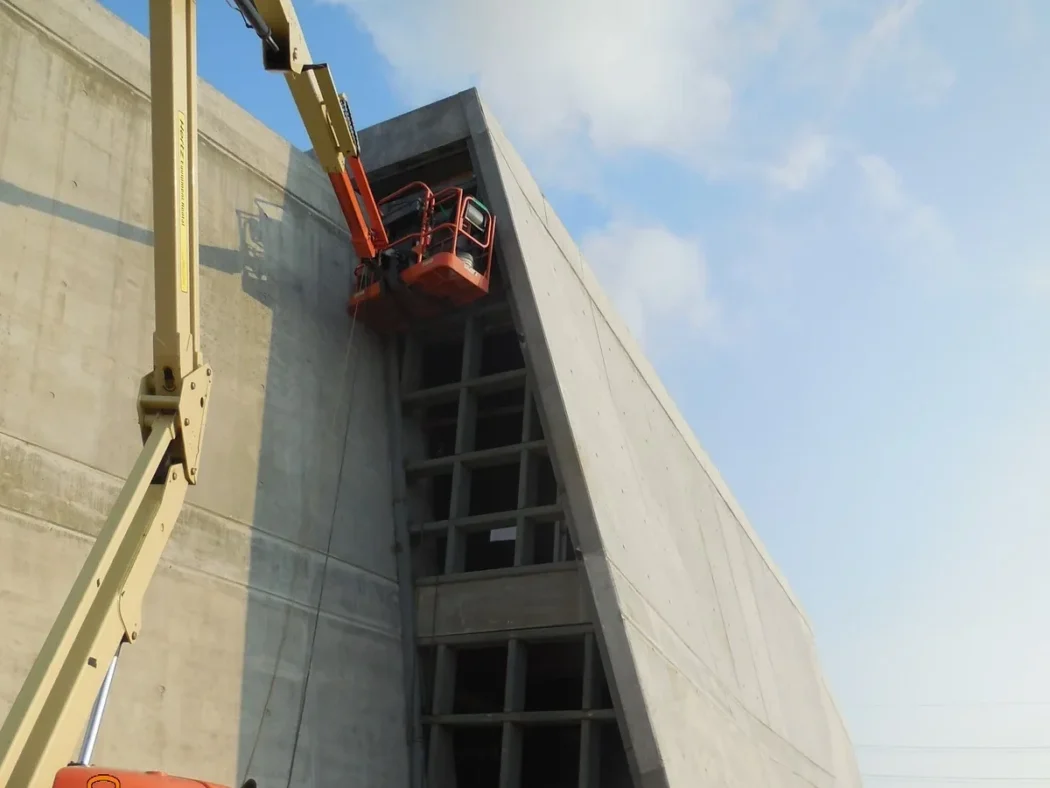A cherry picker lifts a worker in an orange basket to the top of a large, angled concrete structure with rectangular openings on a clear day.