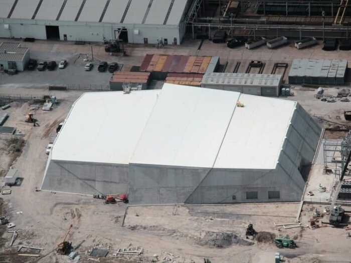 Aerial view of a large, white-roofed, rectangular industrial building under construction, surrounded by equipment and vehicles on a dirt ground.