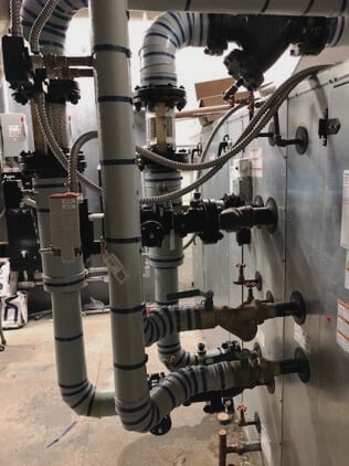 Industrial pipes with blue insulation, part of the Lab HVAC upgrade, connect to a metal panel surrounded by control valves and gauges.