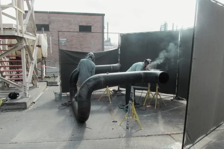 Two workers in helmets are welding large black pipes at a water plant construction site, with protective barriers surrounding the area.