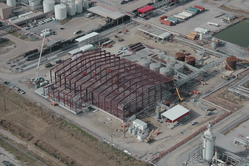 Aerial view of an industrial construction site featuring a large steel frame, warehouse buildings, tanks, and machinery.