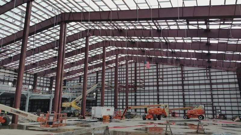 Interior of a large industrial warehouse under construction, with steel beams, manufacturing equipment, and an American flag hanging inside.
