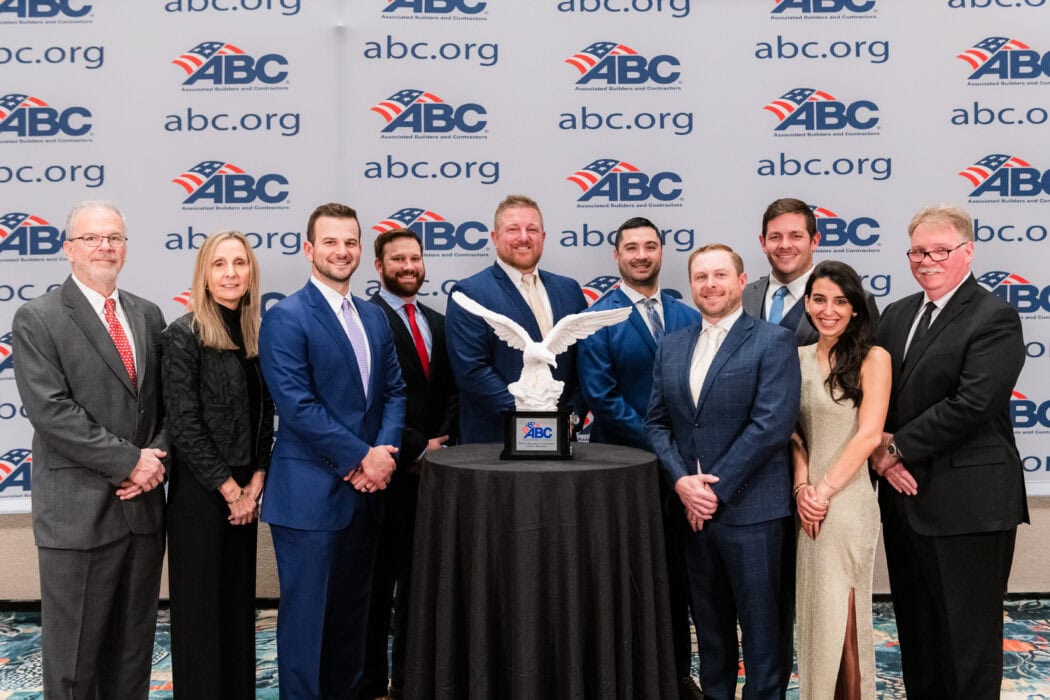 A group of people stand around a table with a trophy in the shape of an eagle. The backdrop displays ABC.org logos.